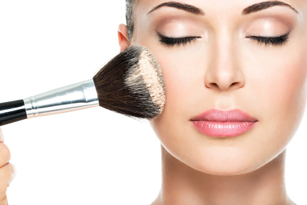 Closeup portrait of a woman applying dry cosmetic tonal foundation on the face using makeup brush.