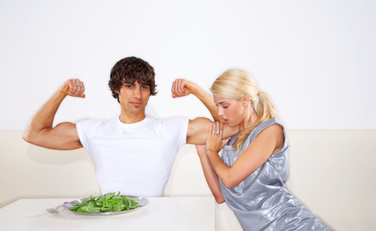 Young man sitting on front of spinach, flexing muscles, woman touching bicep